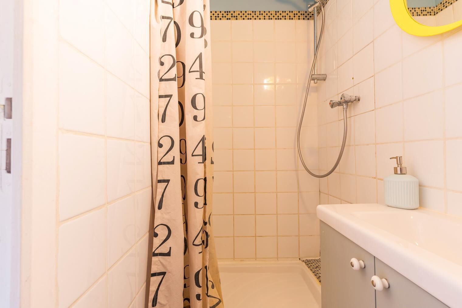 Casa de banho com paredes revestidas a azulejos brancos, chuveiro com área de cerâmica amarela, lavatório branco com dispensador de sabão e porta com padrão numérico.