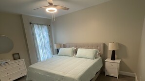 A bedroom with a bed, a nightstand, a lamp, a ceiling fan, and a window with curtains.