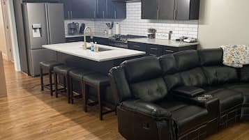 Uma cozinha moderna com armários escuros, uma ilha central e um sofá de couro preto na sala de estar.