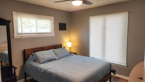Een slaapkamer met een bed, een nachtkastje met lamp, een raam met zonwering, en een plafondventilator.