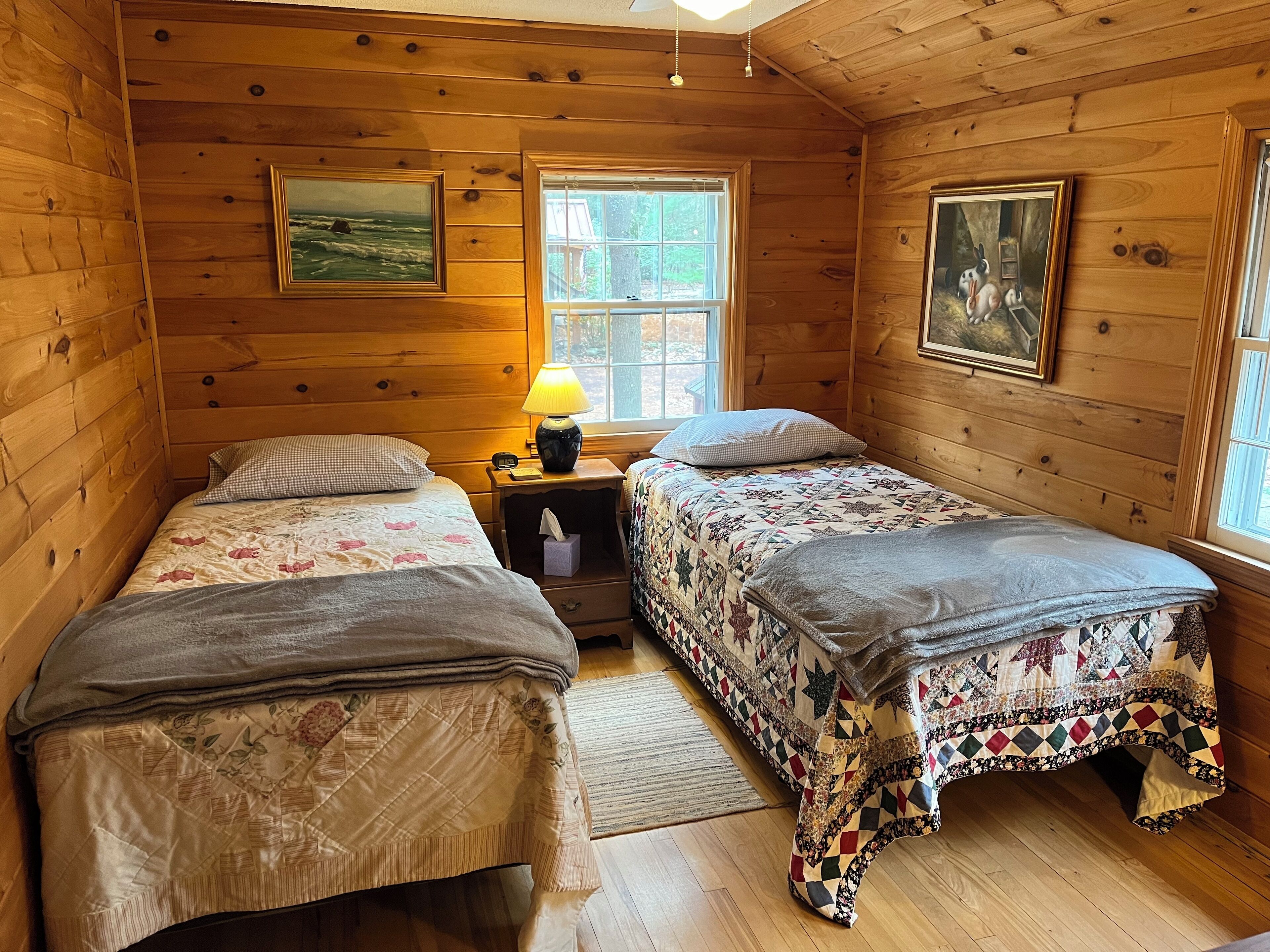 A cabin with two beds, wooden walls, a window, and a lamp.