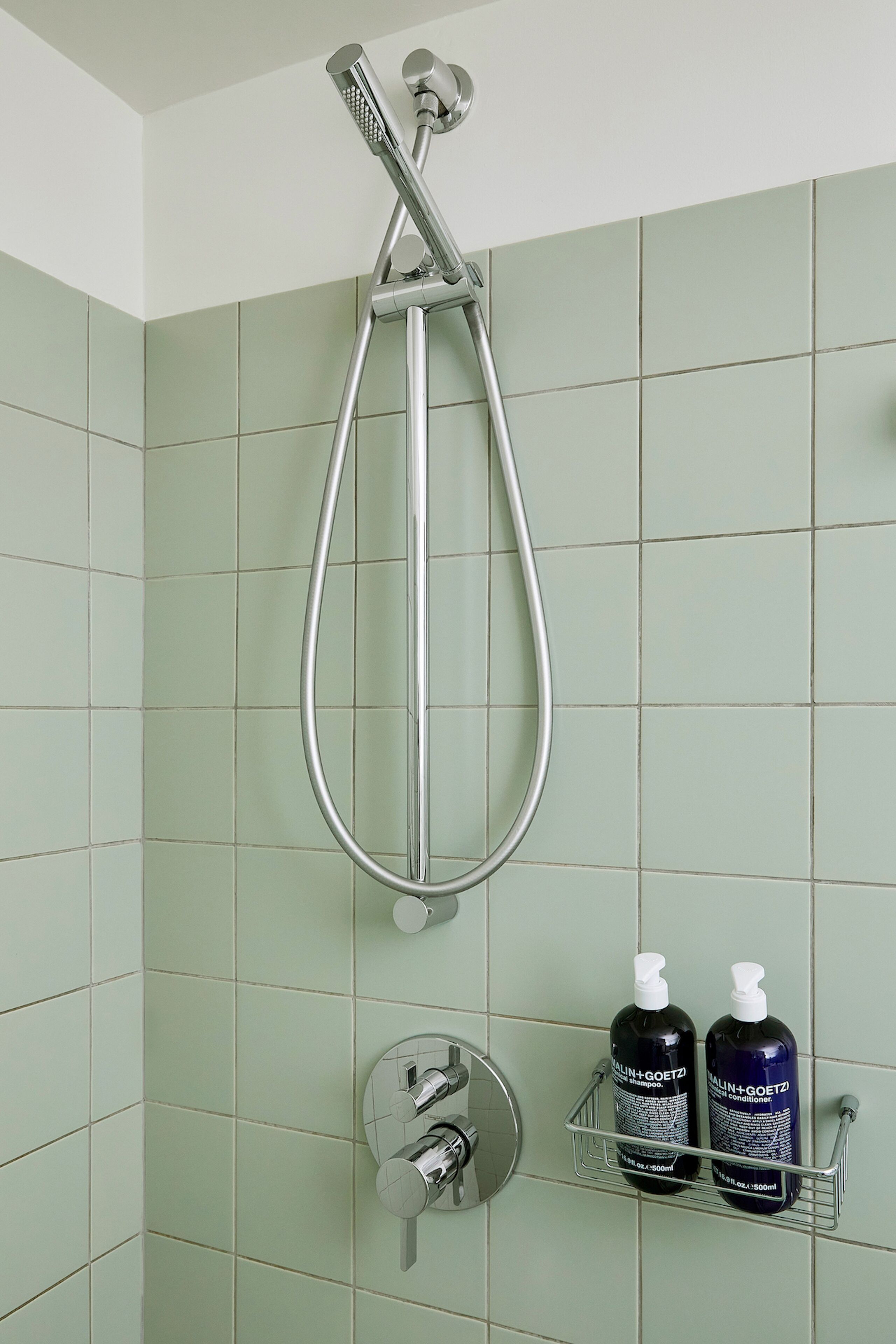 A shower with a handheld showerhead and a wall-mounted faucet. There are two bottles of hair care products on a shelf below.