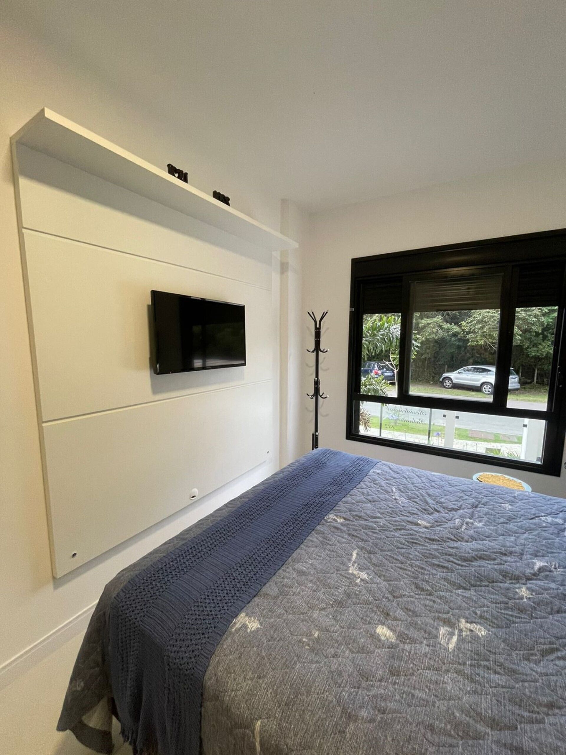 Ein Schlafzimmer mit einem Bett, einem an einer weißen Kommode befestigten Flachbildfernseher, einem Kleiderständer und einem Fenster mit Blick nach außen.