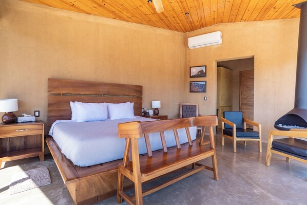 A bedroom with a wooden bed, a bench, two chairs, a nightstand, and an air conditioning unit.