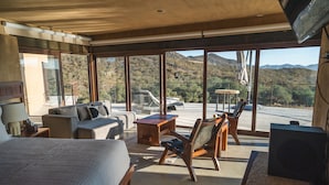 Une chambre d’hôtel moderne avec un grand lit, un canapé, une table basse en bois et un fauteuil. La chambre dispose de grandes fenêtres offrant une vue sur les montagnes.