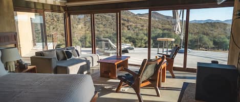 Habitación de hotel moderna con una cama grande, un sofá, una mesa de centro de madera y una silla. La habitación tiene ventanas amplias que ofrecen vistas a las montañas.
