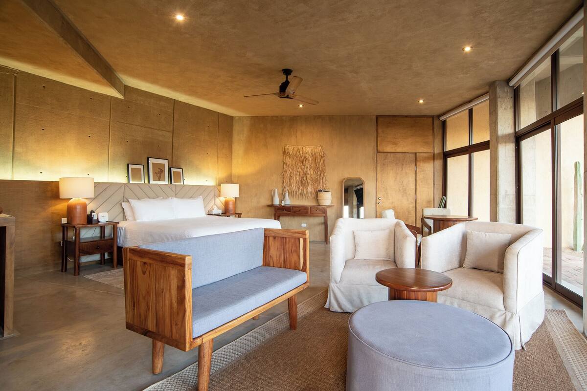 Una habitación de hotel moderna con una cama grande, dos sillones, una mesa redonda y un ventilador de techo.