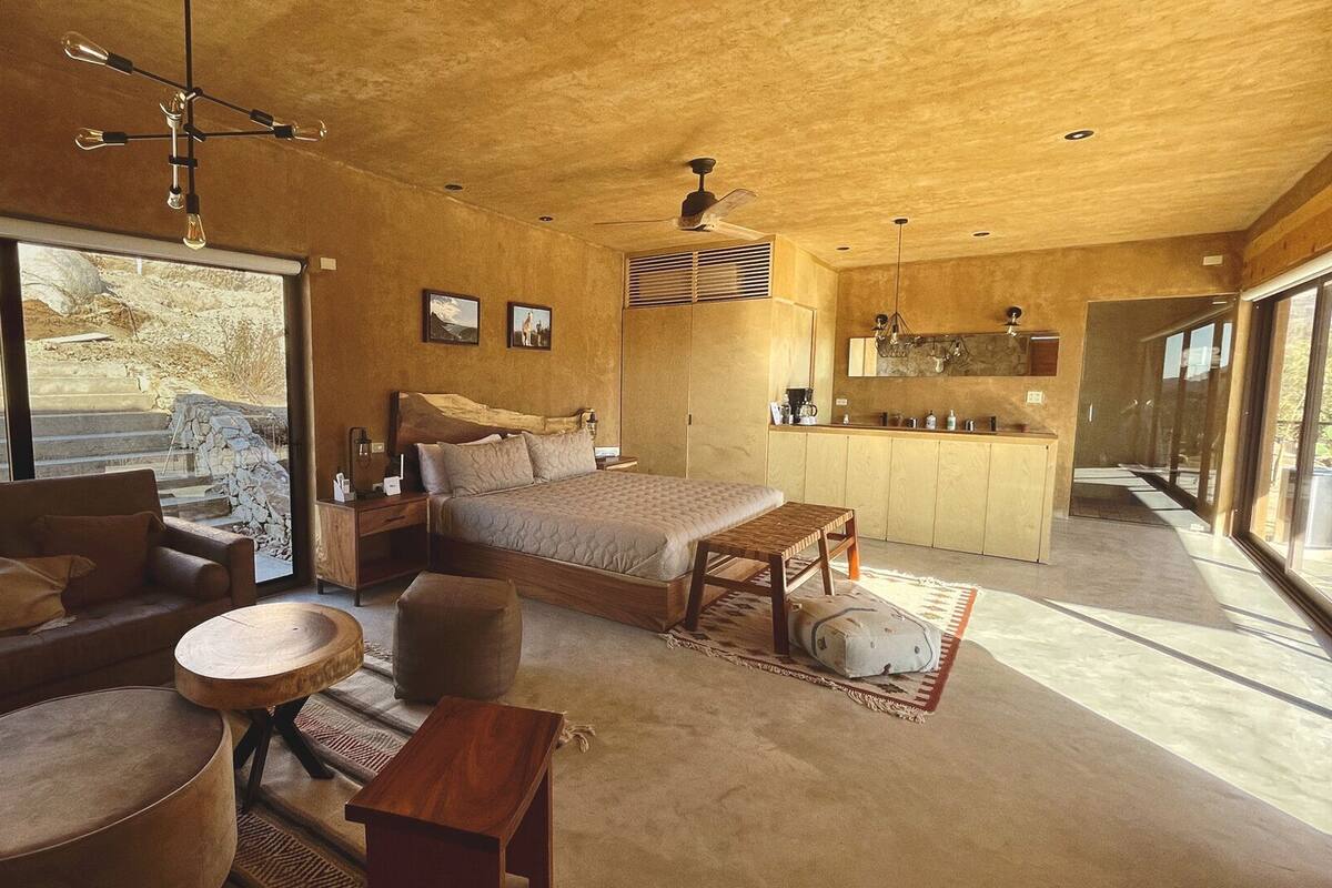 Un interior moderno con una cama grande, sofá y zona de comedor, ventanas amplias y vista al exterior de piedra.