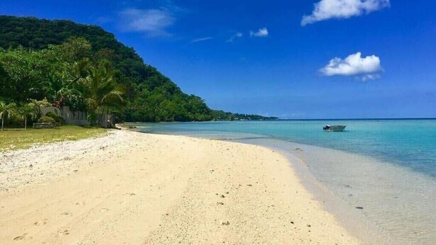 En strand med kristallklart turkost vatten, en sandstrand och ett frodigt, grönt kullområde i bakgrunden.