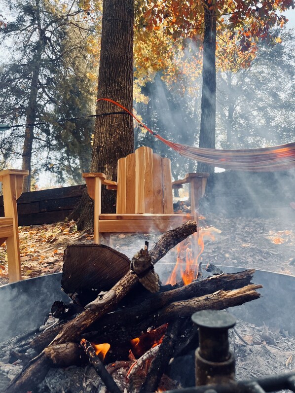 Lejrild med brænde og en metal gryde, omgivet af træstole og en hængekøje i et skovområde.