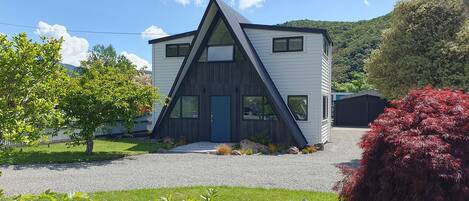 Una casa moderna de dos pisos amb un disseny de teulada triangular, envoltada per un jardí ben cuidat i diversos arbres.