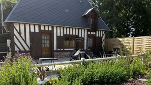 Une maison traditionnelle à colombages, avec un toit en pente, une clôture en bois et un jardin agrémenté de diverses plantes, ainsi qu’une table entourée de chaises.