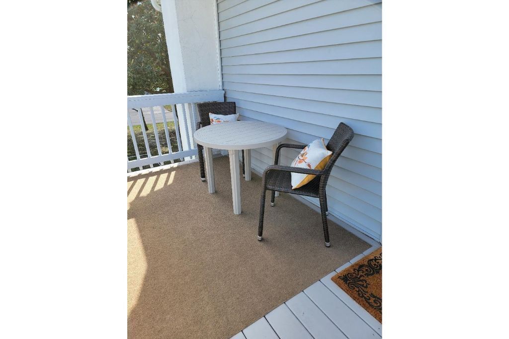 A small outdoor patio with a round table and two chairs, a white railing, and a doormat.