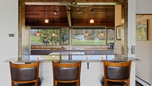 A kitchen with a large island, wooden chairs, pendant lights, and a view of the outdoors.