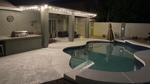 A swimming pool with a curved edge, a covered patio area with string lights, and a barbecue grill. - Free Heated Pool! No pet fee!!  Electric vehicle charger in garage.  (Phoenix)