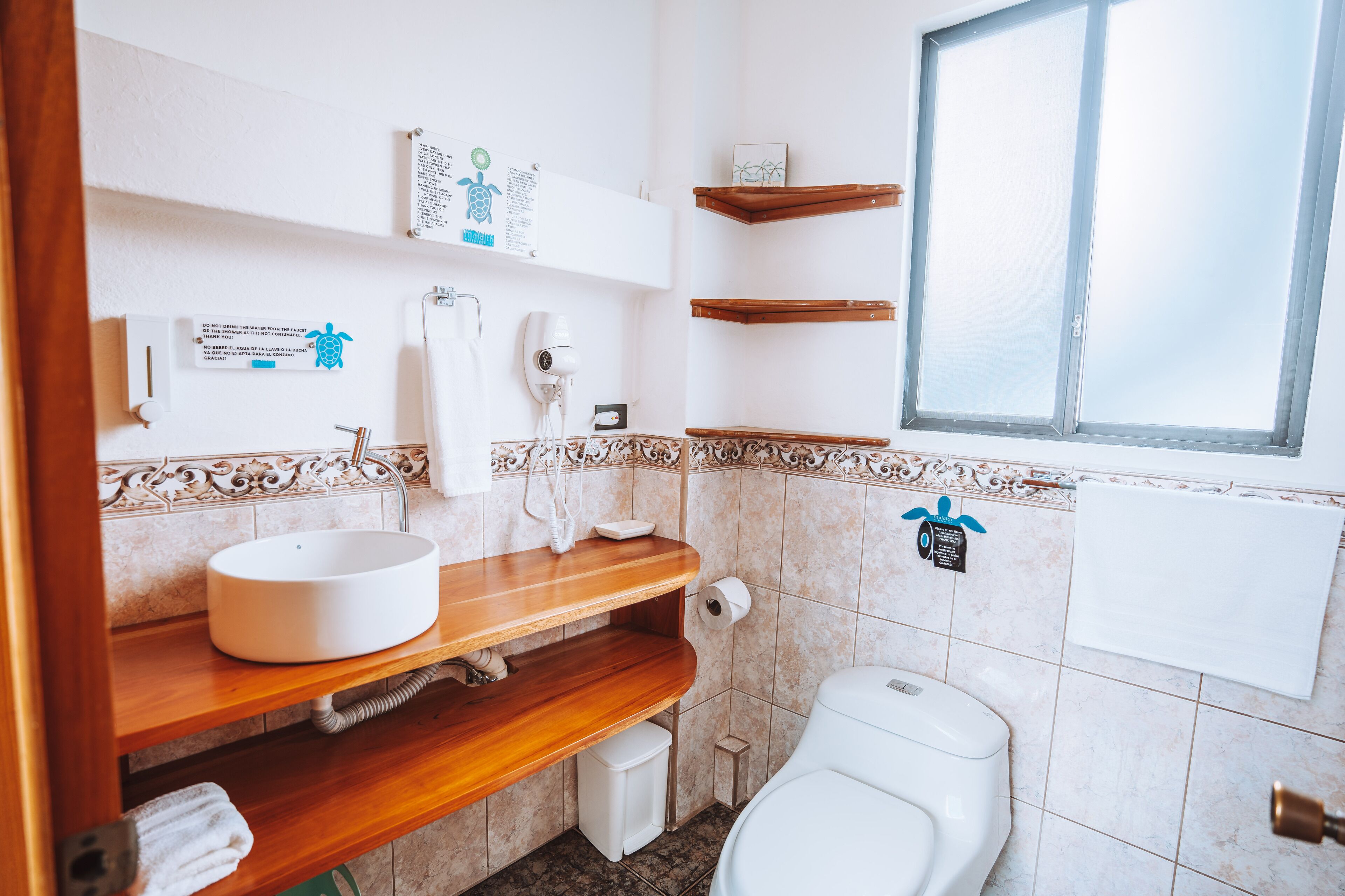 Ein Badezimmer mit einem weißen Waschbecken, einem Holz-Waschtisch und einer Toilette. Über dem Waschbecken befinden sich Regale und rechts ist ein Fenster.