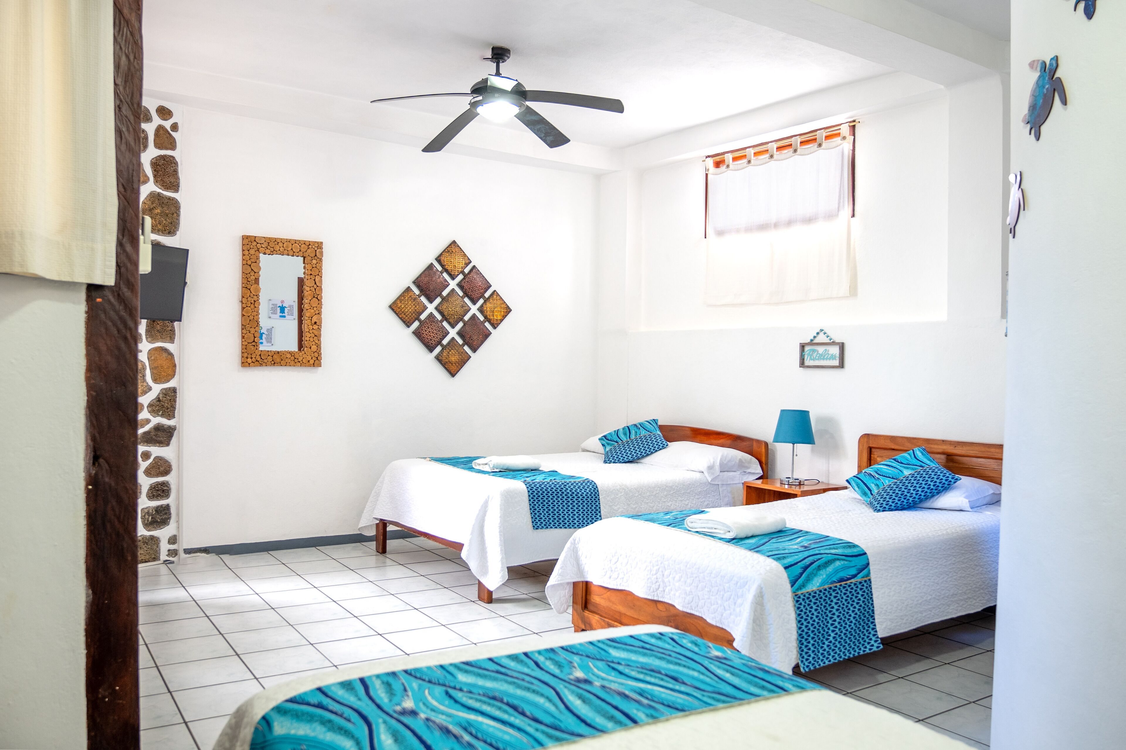 Habitación de hotel con dos camas, ventilador de techo, ventana con cortinas y pared de piedra.