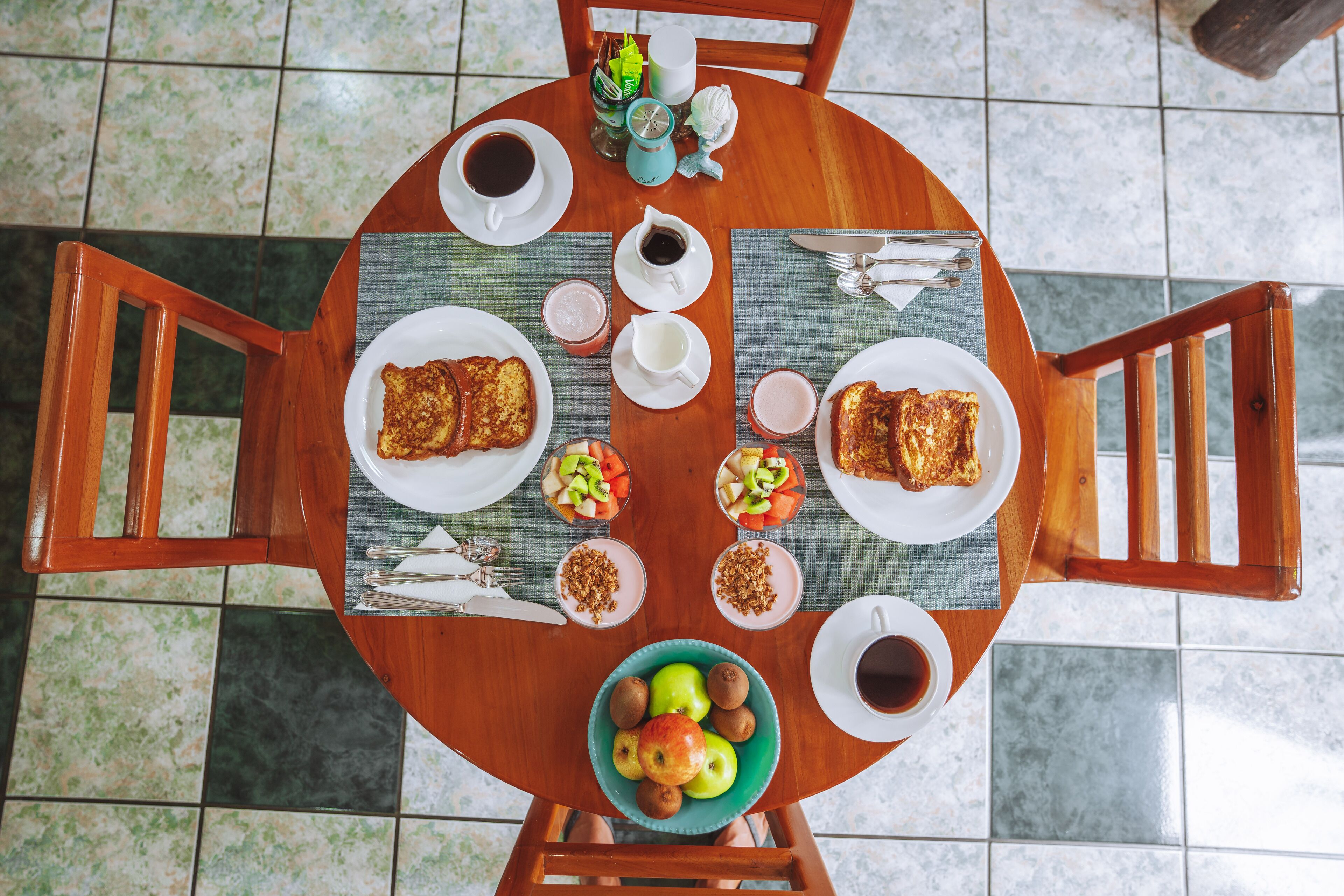 A round wooden table with breakfast items including toast, fruit, and beverages, surrounded by chairs.