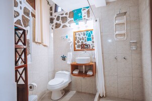 Un baño con inodoro, lavabo y ducha. Hay un perchero y un espejo sobre el lavabo.