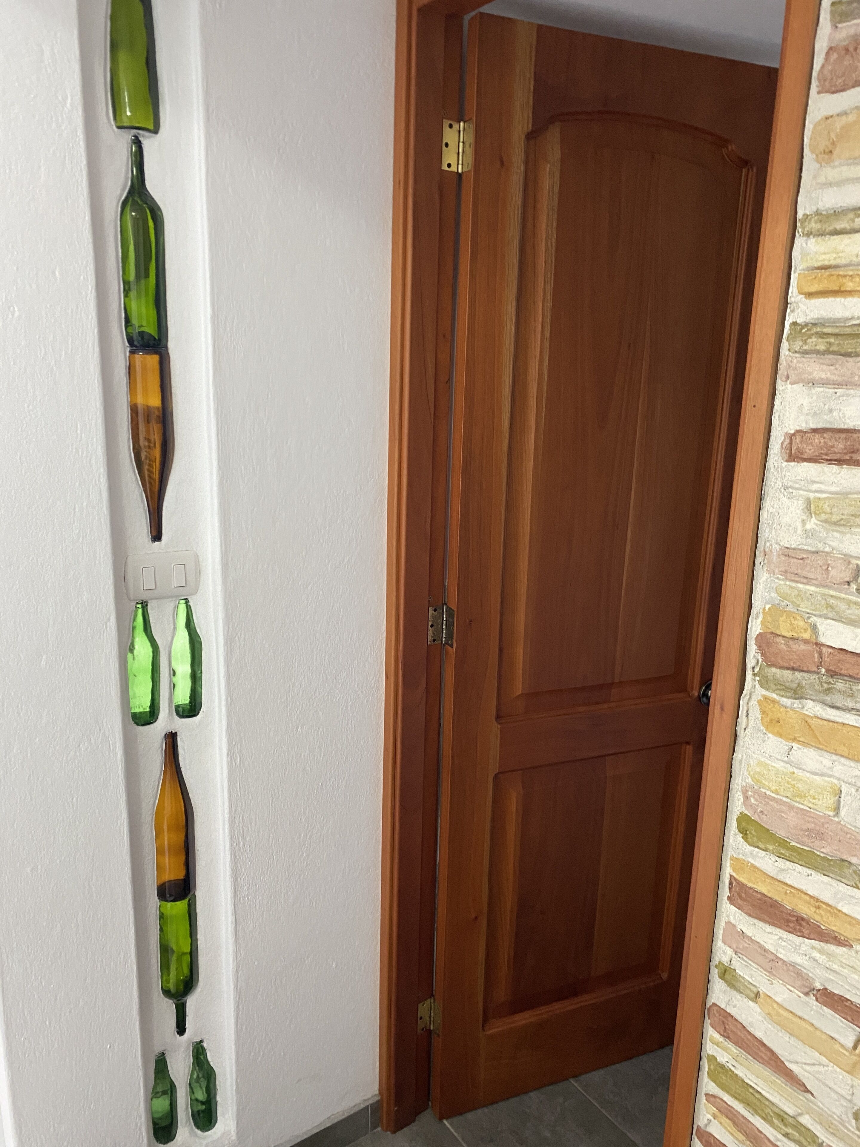 A wooden door with a metal latch, a white wall with a vertical arrangement of green glass bottles, and a stone-patterned wall to the right.