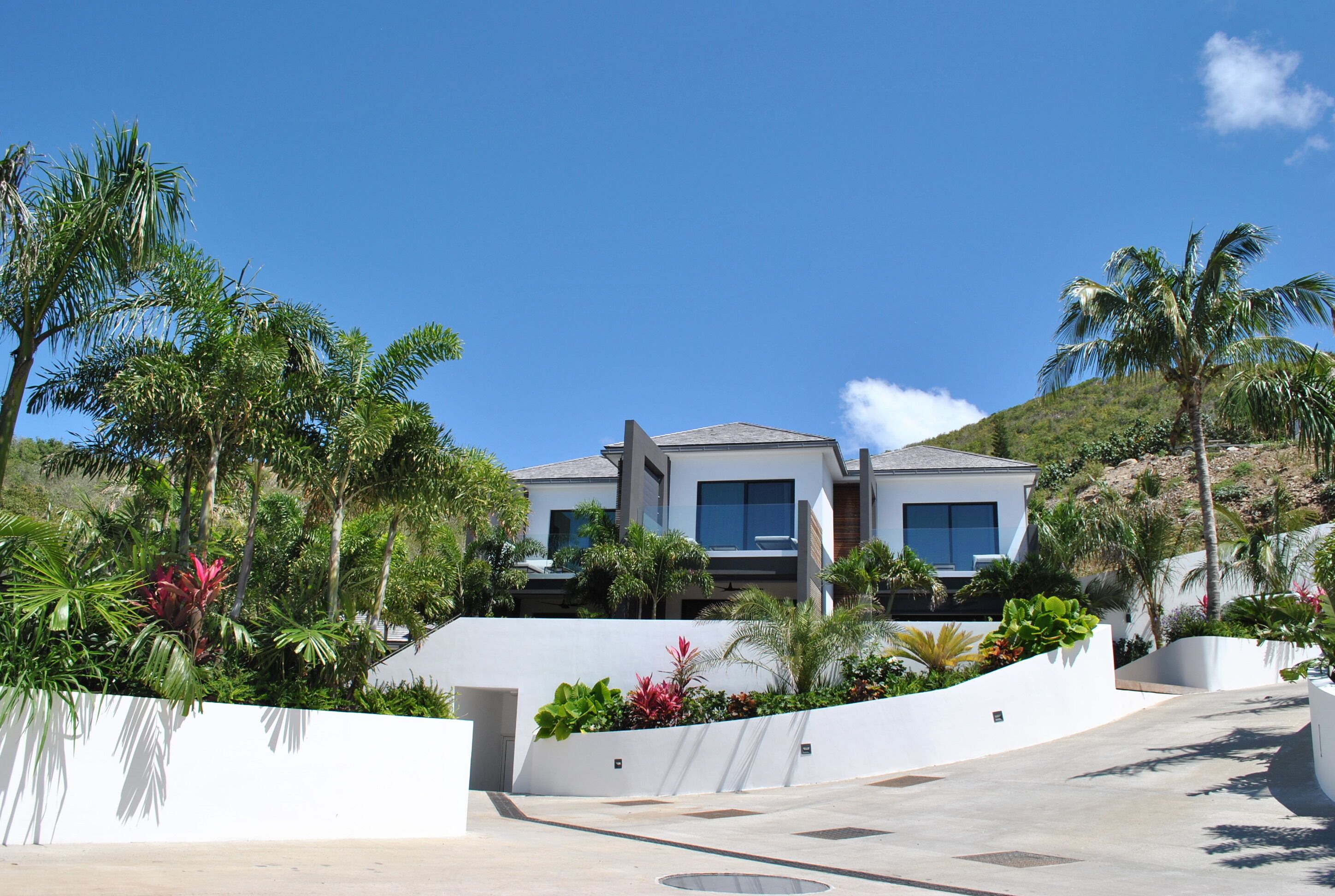 A modern two-story house with a white exterior, surrounded by lush greenery and palm trees.