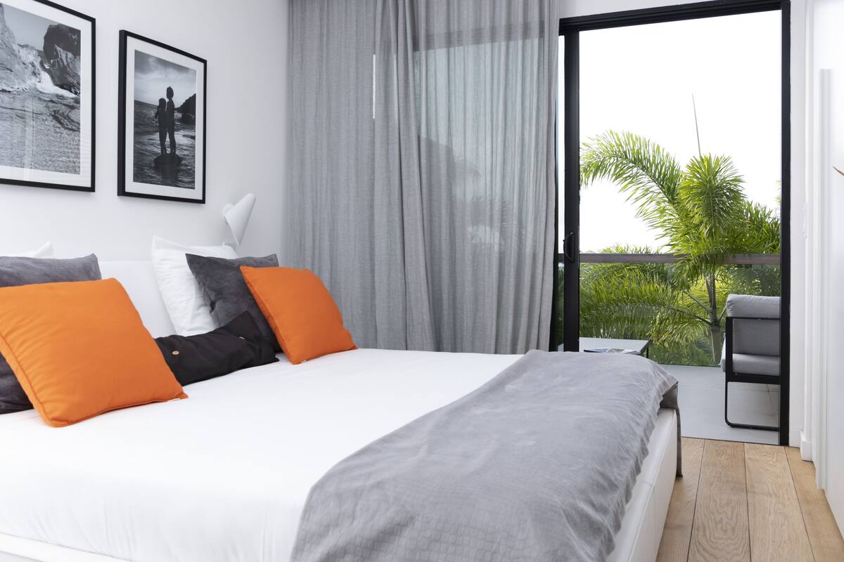 A bedroom with a large bed, grey and orange pillows, a grey blanket, and a view of greenery through a sliding glass door.