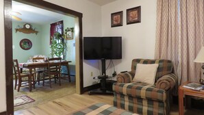A living room with a television, a plaid armchair, a sofa, a dining area with a table and chairs, and a clock on the wall.