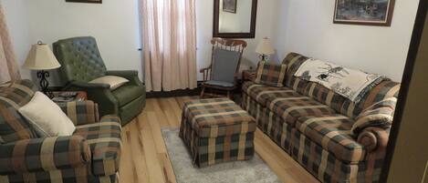 A living room with a green armchair, a sofa, and a plaid-patterned ottoman. There are two framed pictures on the walls and a window with curtains.