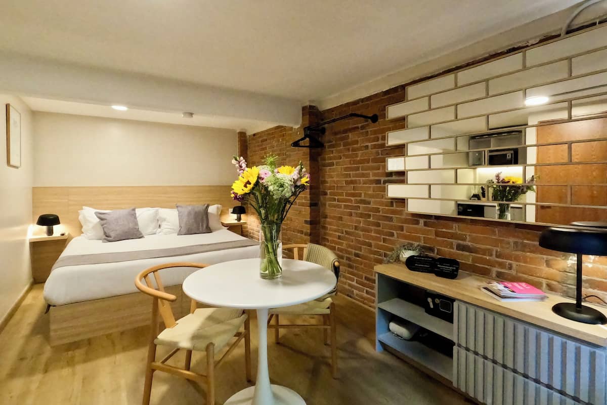 Una habitación de hotel con una cama, una mesa redonda con un florero, una silla, un aparador con espejo y un ventilador de techo.