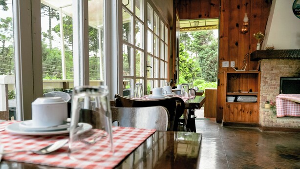 A dining area with a table set for two, large windows, and a fireplace. - Pousada Casa de Campos Capivari by Concavus (Campos do Jordão)