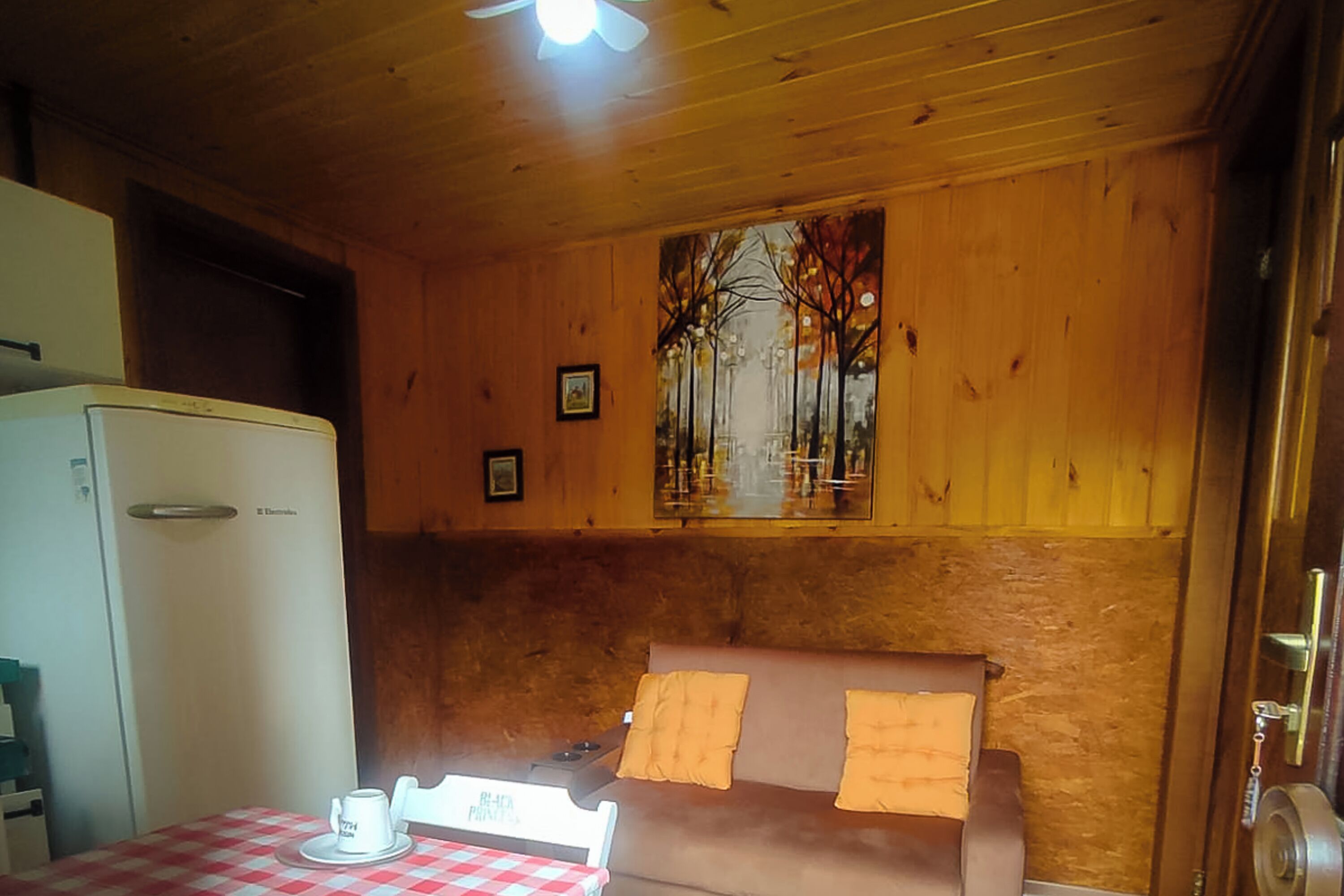 A cozy room with a wooden ceiling, a dining table with a checkered tablecloth, a sofa with cushions, a refrigerator, and a painting on the wall.