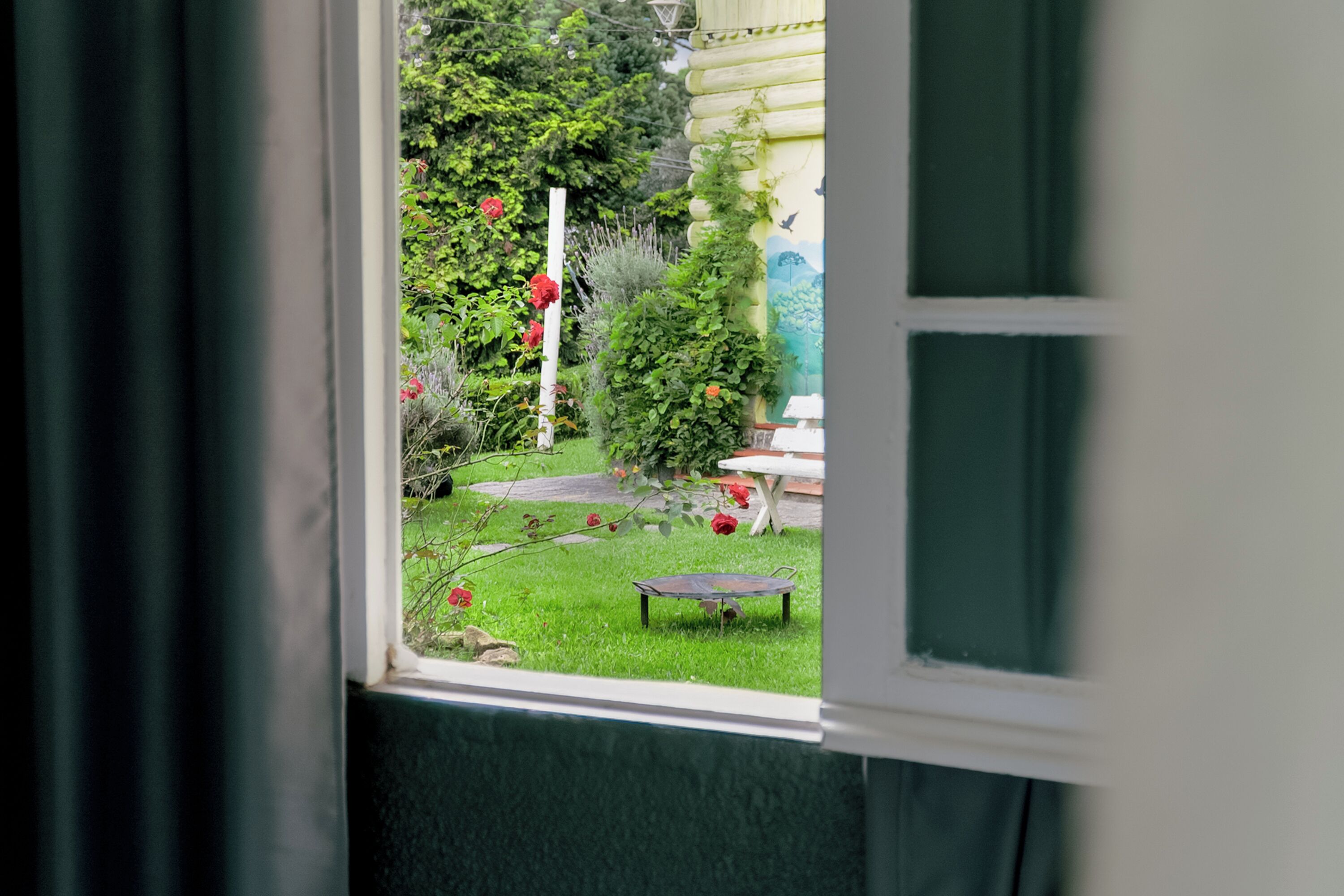 A window view of a garden with a white table, a black table, and red flowers.