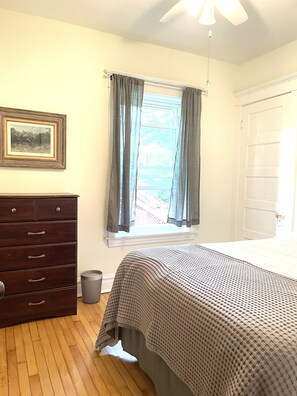A bedroom with a wooden dresser, a window with curtains, a bed with a checkered blanket, and a ceiling fan. - Clean and Comfy Updated Apt in Convenient Location (Forest Park)