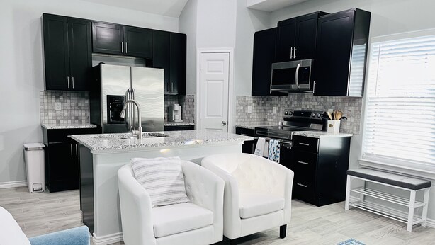 A modern kitchen with black cabinets, stainless steel appliances, and a white island. There is a white sofa and a blue chair in the living area. - Beautiful home 5 minutes to Lake, water activities, shopping and restaurants (Montgomery)