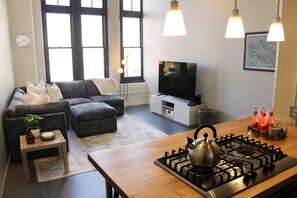 Ein modernes Wohnzimmer mit einer Couch, einem Fernseher, einer Küchenzeile mit Gasherd und einem Esstisch.