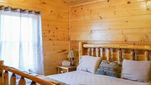 Una habitación tipo cabaña de madera con una cama, una mesita de noche y una ventana con cortinas.