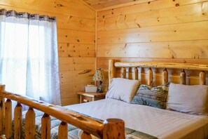 Una habitación tipo cabaña de madera con una cama, una mesita de noche y una ventana con cortinas.