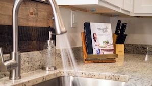 Een keuken met een spoelbak van roestvrij staal, een kraan, een glazen vaas en een houten standaard waarop het boek 'Magnolia Table' staat.