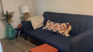 A cozy living room with a dark blue sofa, a small orange ottoman, a lamp, and a vase with flowers on a side table.