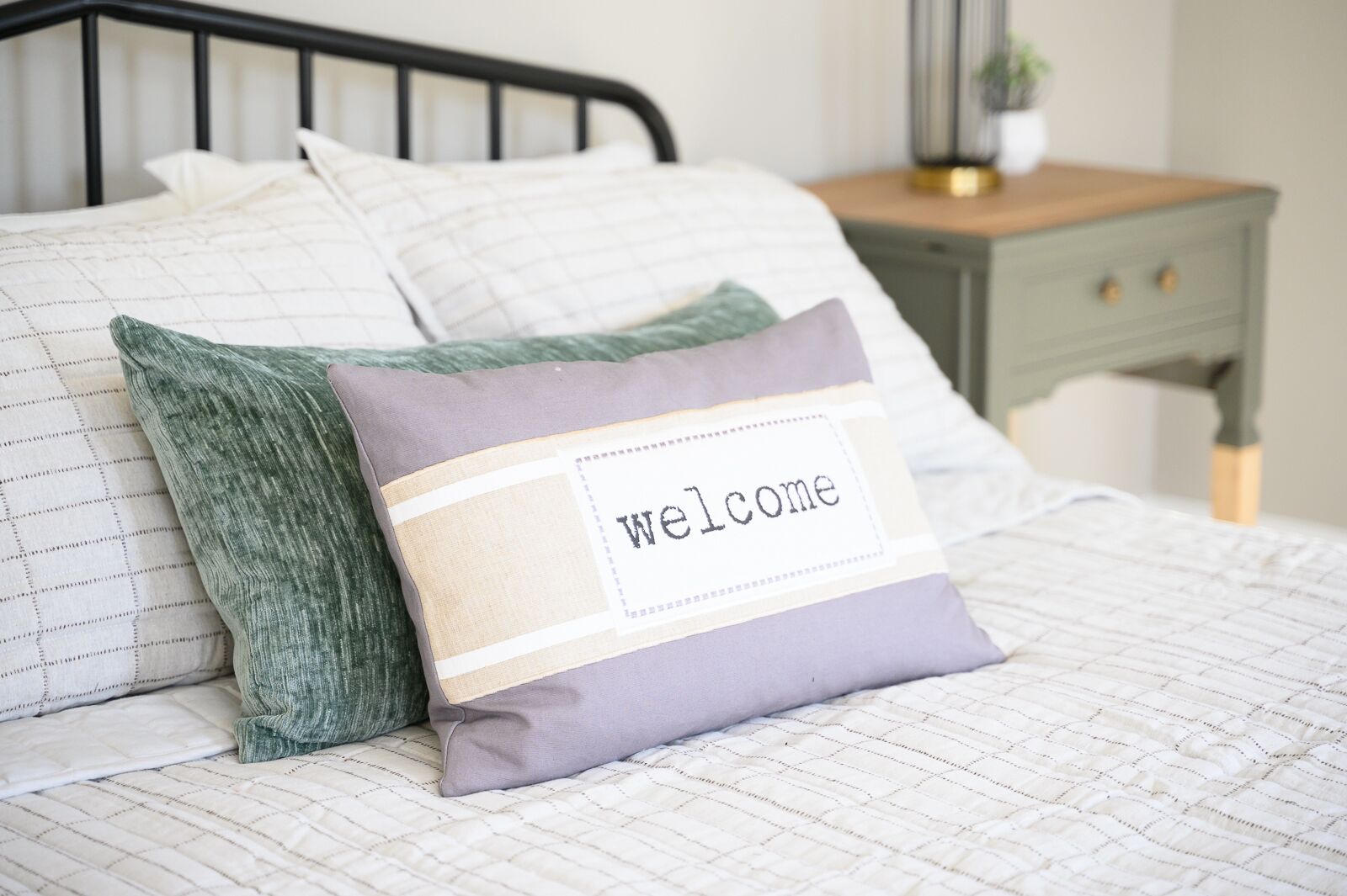 Un lit avec une couette et des draps blancs et gris matelassés, un oreiller vert et un oreiller violet avec une étiquette indiquant «Bienvenue».