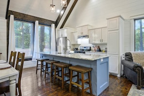 A kitchen with white cabinets, a large island with stools, a dining table with chairs, and a view of trees through the windows. - -Stay A Spell Cabin- (Broken Bow)