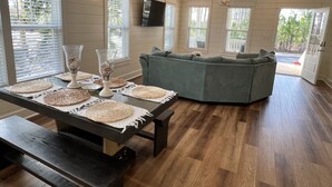 Un salon moderne avec une table en bois foncé, des sets de table en fibres tressées, un canapé d’angle gris et de grandes fenêtres équipées de stores.