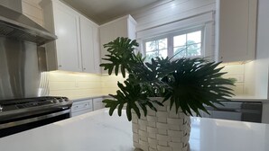 Une cuisine avec des armoires blanches, une hotte de cuisinière en acier inoxydable et un îlot central blanc agrémenté d’un pot de fleurs en osier.