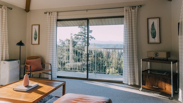 A room with a large sliding glass door leading to a balcony with a mountain view. There is a wooden table with a book and a vase, a chair with a cushion, and a small cabinet with a radio. - Iconic Union Skyhouse with Hood Canal views (Union)