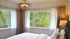 A bedroom with a large bed, two windows with curtains, a chandelier, and a view of greenery outside. - Iconic Union Skyhouse with Hood Canal views (Union)