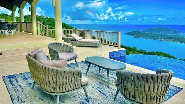Un área de estar al aire libre cubierta con sillones reclinables y una mesa, con vistas a una piscina y un paisaje pintoresco del mar y las colinas.