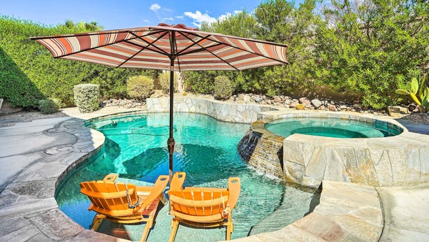 A swimming pool with two wooden chairs and a striped umbrella. - Indio Home w/ Private Pool & Community Amenities (Indio)