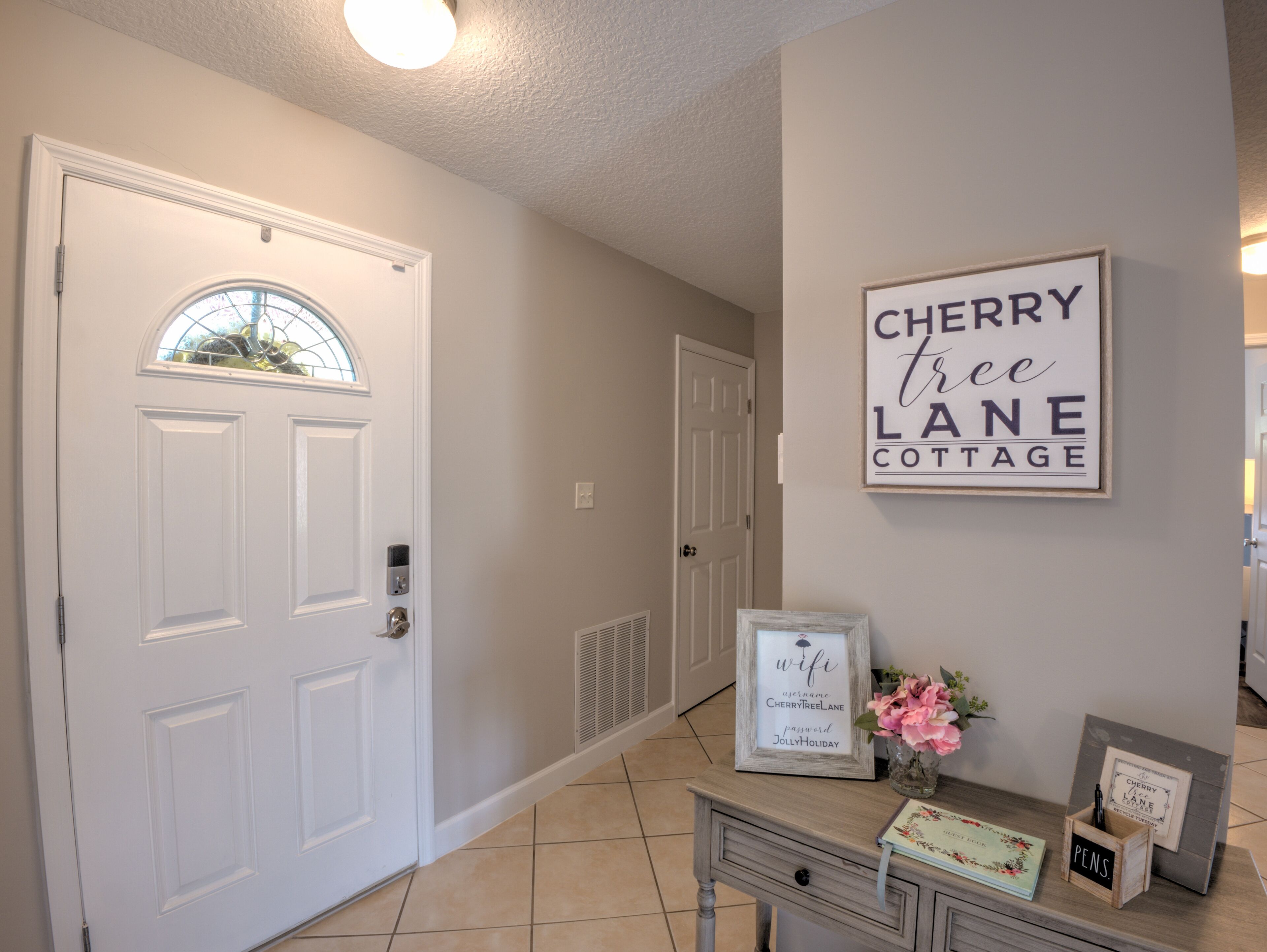 A hallway with a white door, a sign reading 'Cherry Free Lane Cottage', and a small table with a vase and framed items.