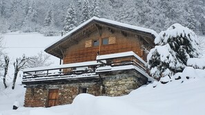 Une cabane en bois, posée sur des fondations en pierre, entourée d’arbres couverts de neige.