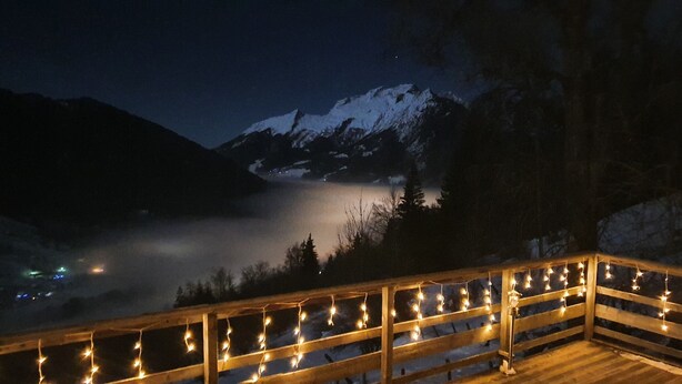 A wooden deck with string lights, overlooking a snow-covered mountain and a valley with a foggy lake.
